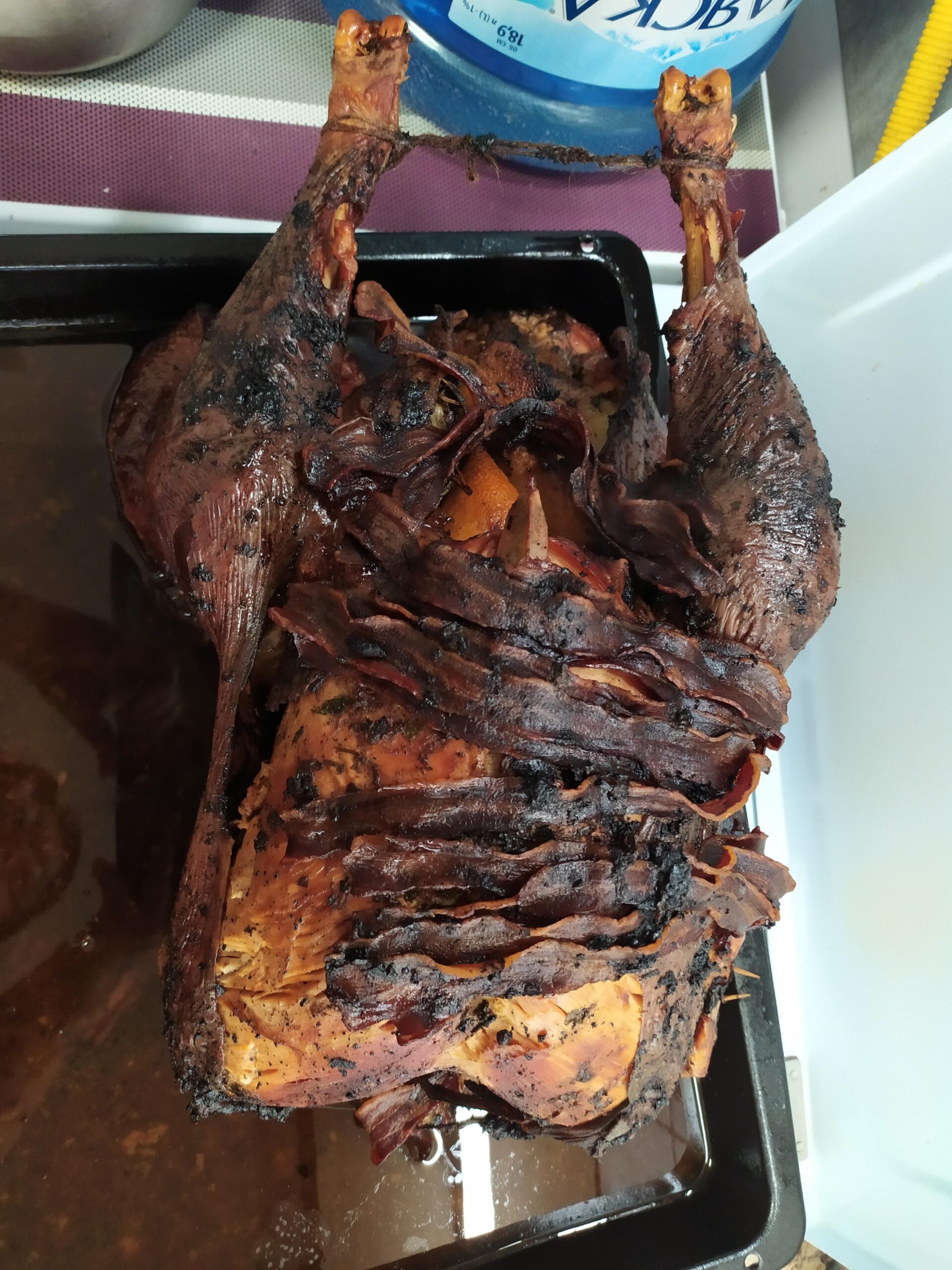 A whole roasted bird, possibly turkey or chicken, with crispy, dark bacon strips on top, lies in a black roasting pan. Thankfully, this culinary triumph avoids any hilarious cooking disasters—the bird appears well-cooked with visible herbs and perfectly browned skin.