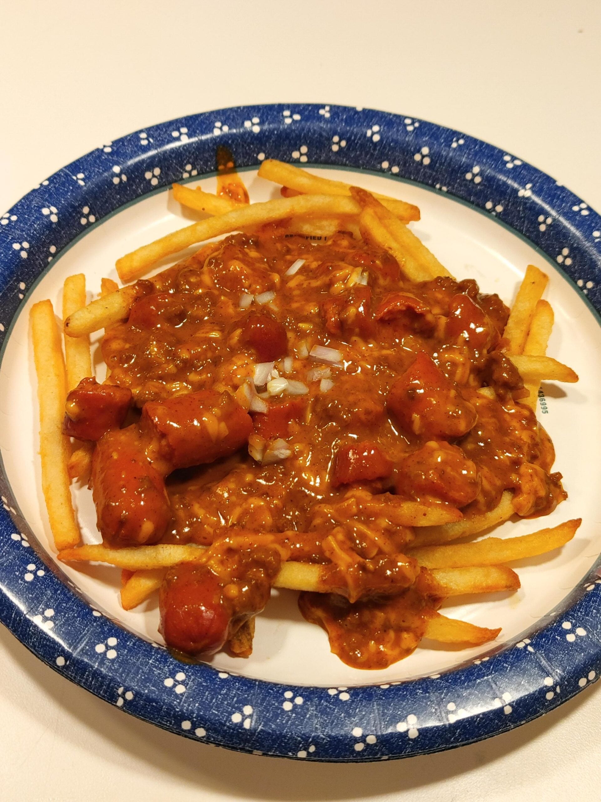 French fries topped with chili, chopped onions, and melted cheese on a blue and white paper plate.