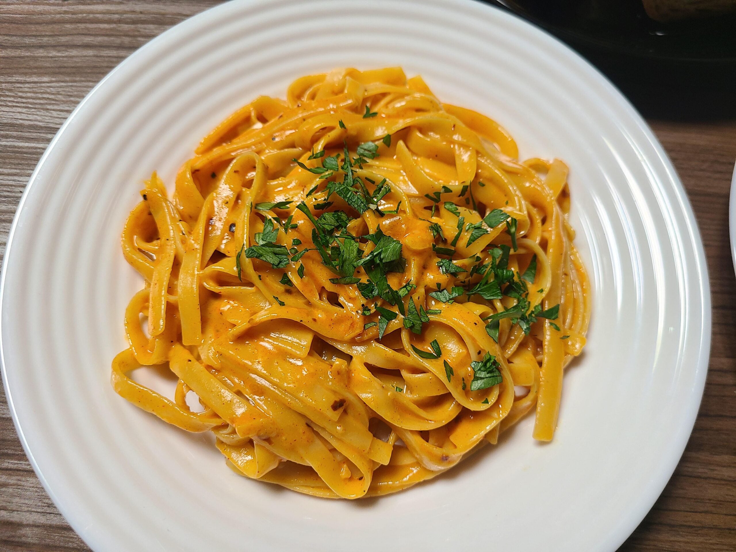 A white plate of fettuccine pasta in a creamy orange sauce, garnished with chopped fresh parsley, sits on a wooden table.
