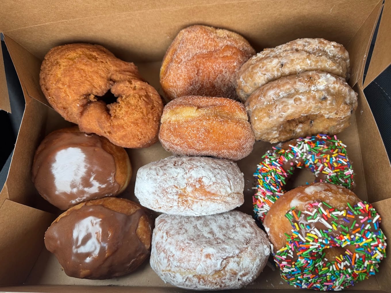 A box of assorted donuts including powdered, chocolate-glazed, sugar-coated, cinnamon, old-fashioned, and one with colorful sprinkles. The donuts are arranged in three rows inside a brown box.