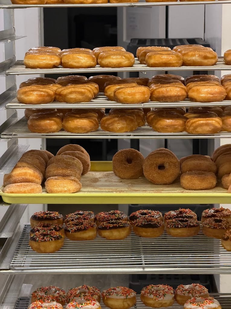 Metal racks filled with trays of assorted donuts, including glazed, sugar-coated, and donuts with chocolate frosting and colorful sprinkles, displayed in a bakery setting.