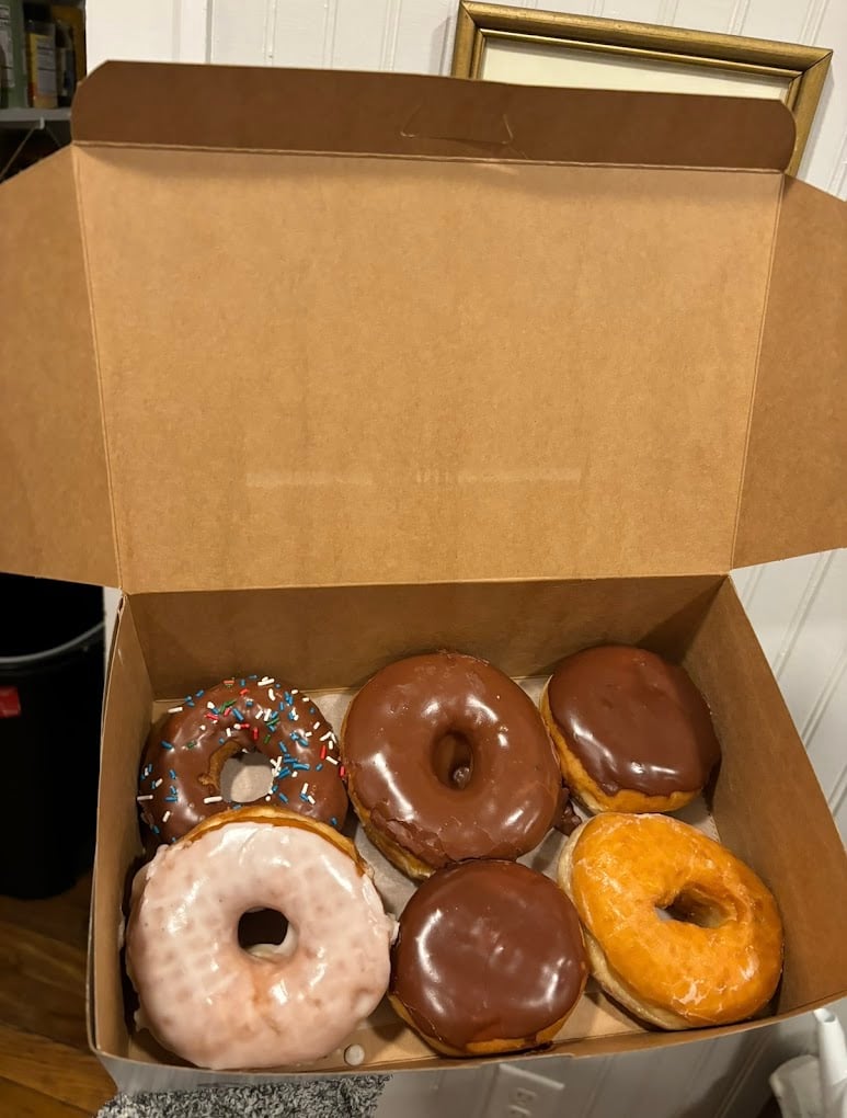 An open box containing six donuts: three chocolate glazed, one with chocolate glaze and sprinkles, one plain glazed, and one with white glaze, all resting in a brown cardboard box.