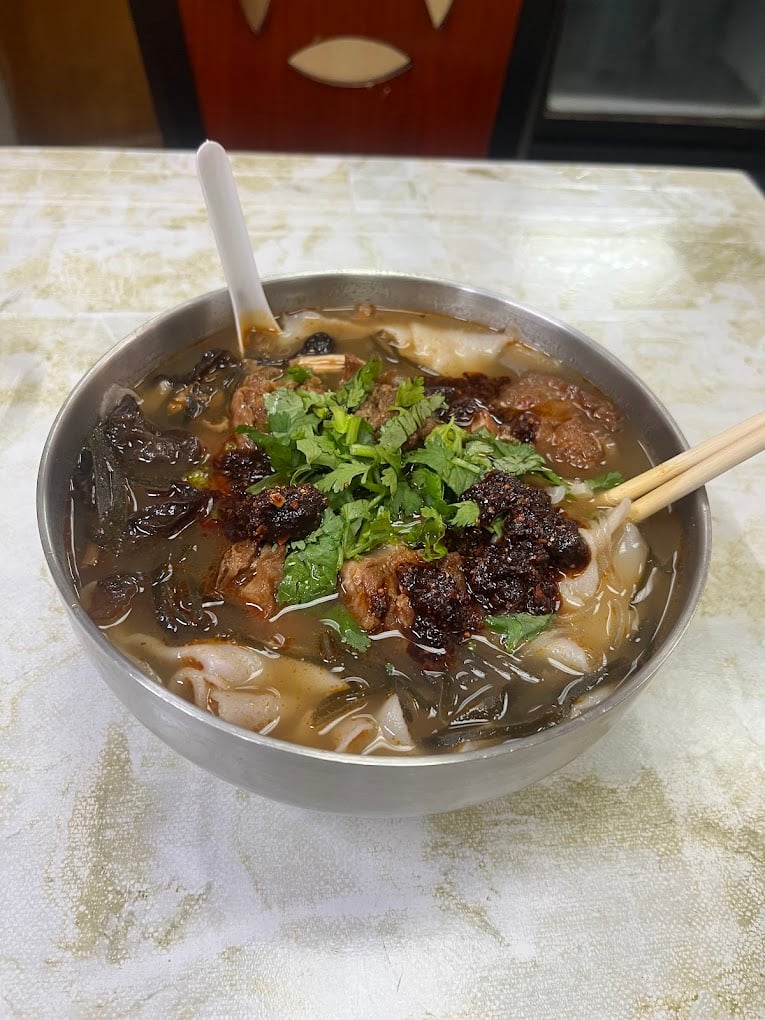 A bowl of noodle soup topped with fresh cilantro, chili paste, wood ear mushrooms, and slices of meat, with a spoon and chopsticks resting on the bowl.