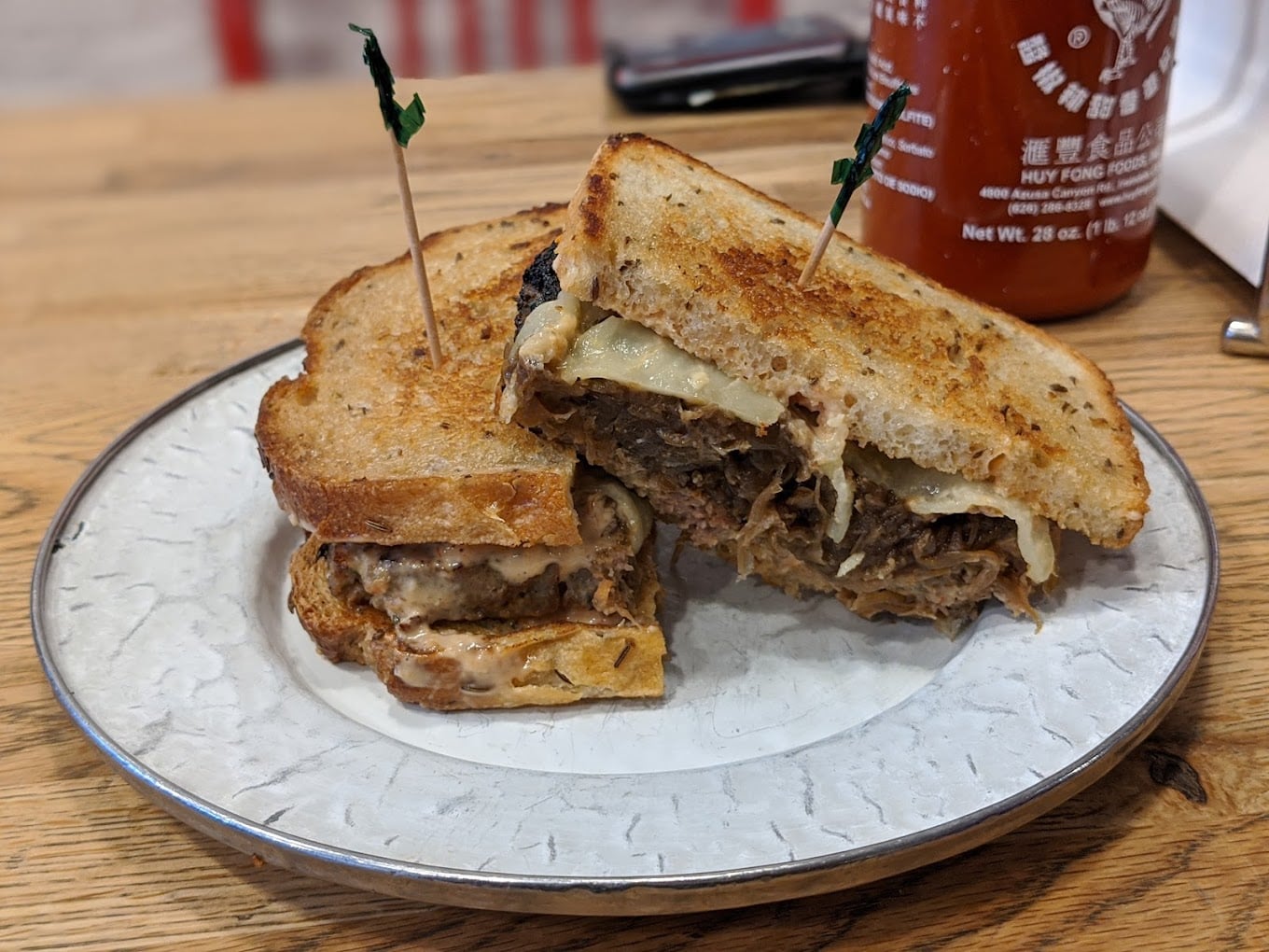 A grilled sandwich filled with shredded meat and melted cheese, cut in half and held together with toothpicks, is served on a white plate. A bottle of sriracha sauce is visible in the background.
