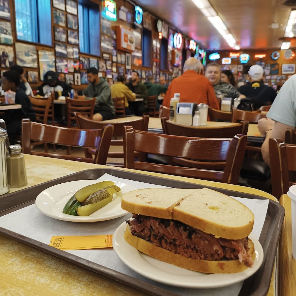 A deli sandwich filled with sliced meat and served with pickles sits on a tray atop a table in a bustling, vintage-style restaurant filled with customers and decorated walls.