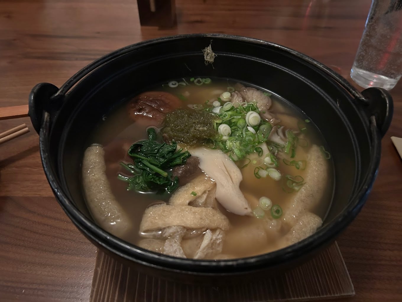 A bowl of noodle soup with sliced mushrooms, chopped green onions, leafy greens, tofu, and seaweed in a clear broth, served in a black pot on a wooden table.
