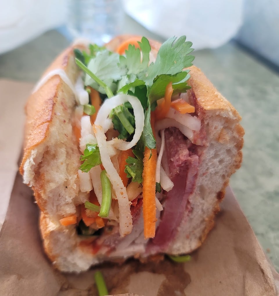 A close-up of a banh mi sandwich on a brown paper wrapper, filled with sliced meats, pickled carrots, daikon, cucumber, fresh cilantro, and jalapeño on a crusty baguette.