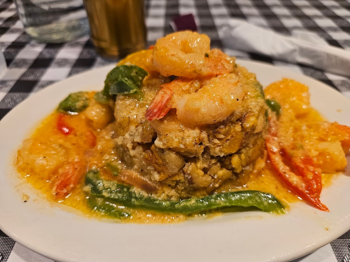 A plate of mofongo topped with shrimp in a creamy, yellow sauce, garnished with red and green peppers, served on a white dish over a black and white checkered tablecloth.