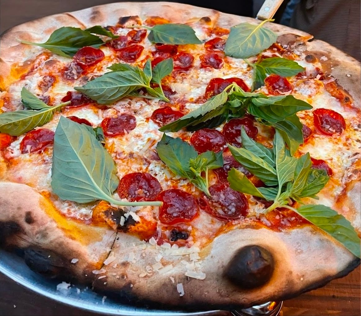 A wood-fired pepperoni pizza topped with melted cheese and fresh basil leaves, with a slightly charred, crispy crust, served on a metal tray.