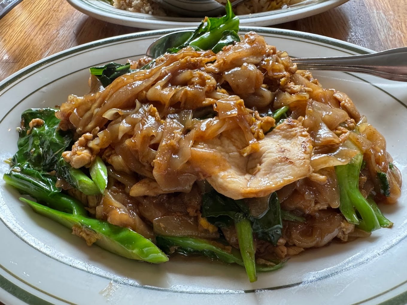 A plate of stir-fried flat rice noodles with sliced pork, egg, and Chinese broccoli in a savory sauce, served on a white dish with a spoon.