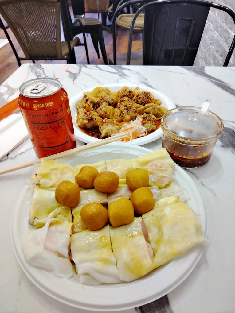 A marble table with plates of dim sum: rice noodle rolls topped with fish balls, dumplings with sauce and shredded vegetables, a small cup of chili oil, and a can of Sapporo soda.
