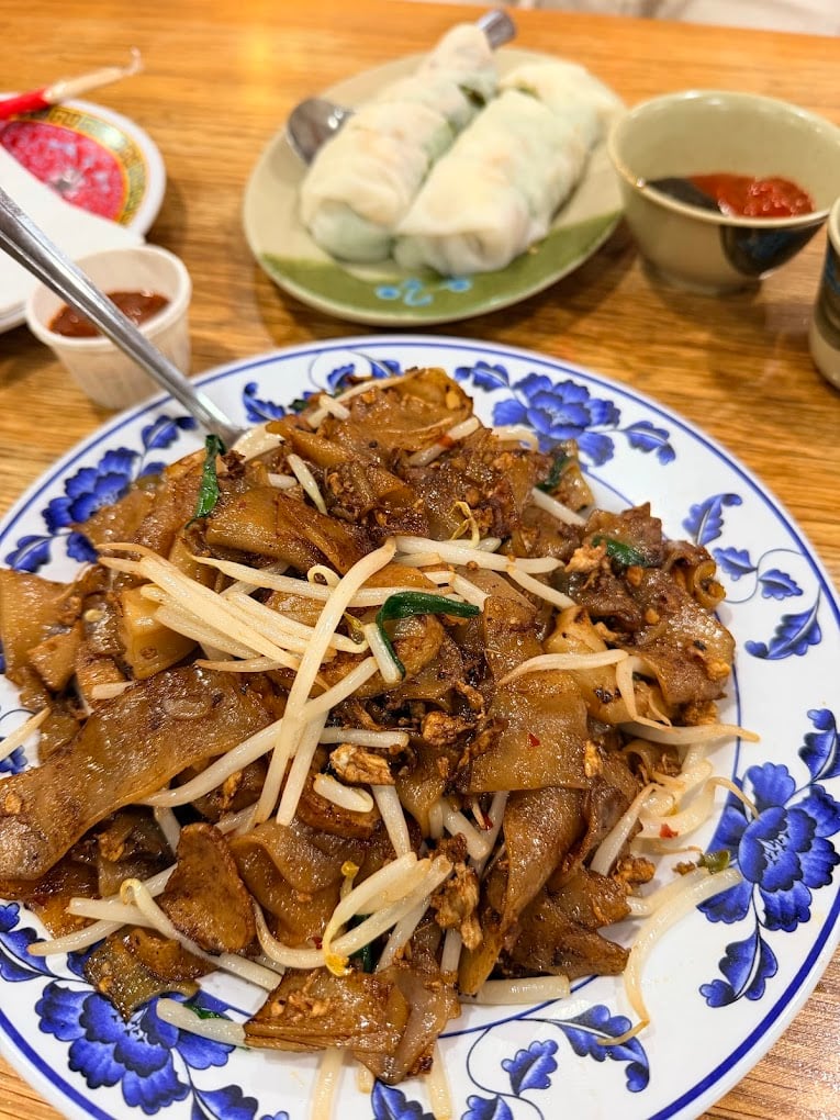 A plate of stir-fried flat noodles with bean sprouts and egg sits on a blue and white dish, with fresh spring rolls and dipping sauces in the background on a wooden table.