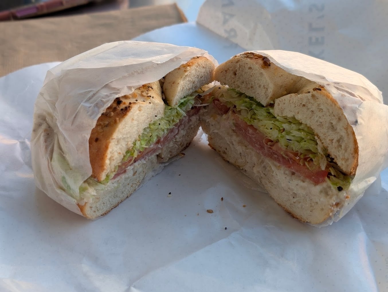 A sliced bagel sandwich with lettuce, tomato, and what appears to be a spread, wrapped partially in white paper and placed on white deli paper.