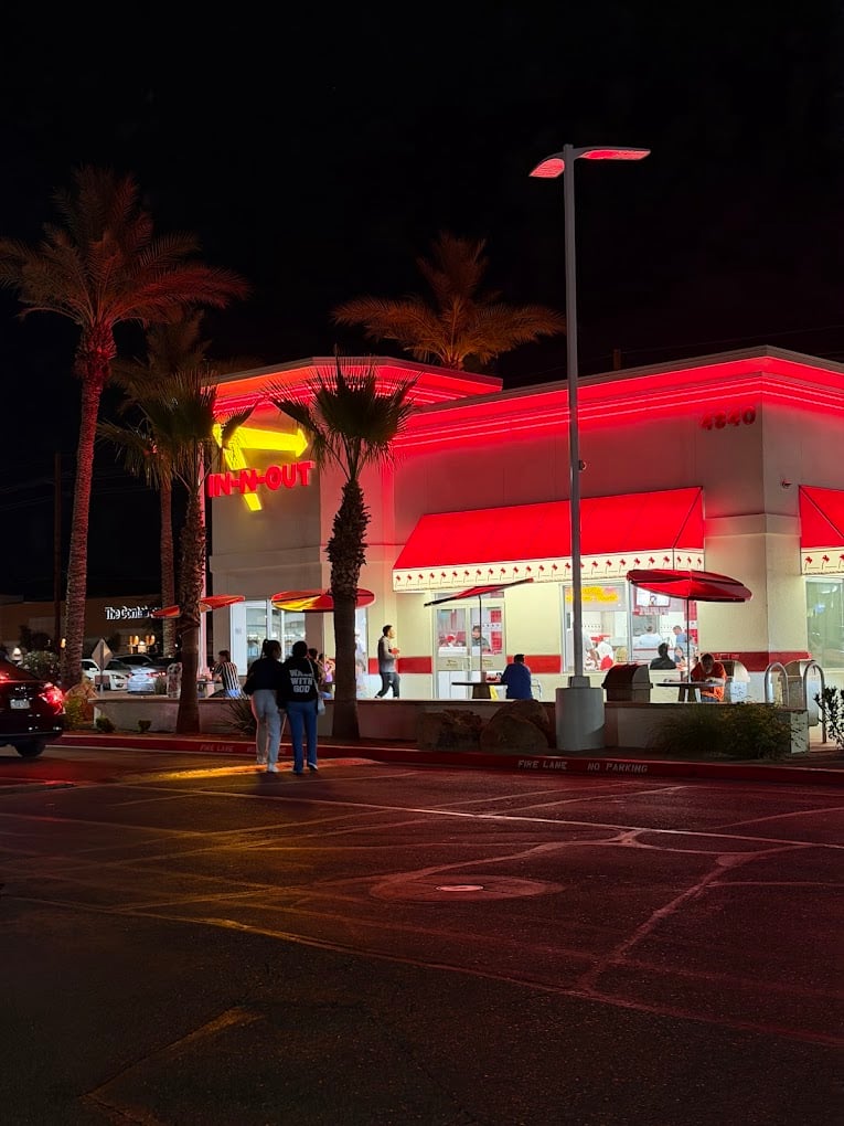 A brightly lit In-N-Out Burger restaurant at night, with people standing outside and palm trees nearby. As one of the favorite fast-food chains by state, its building glows with red neon lights, and cars are parked in front.