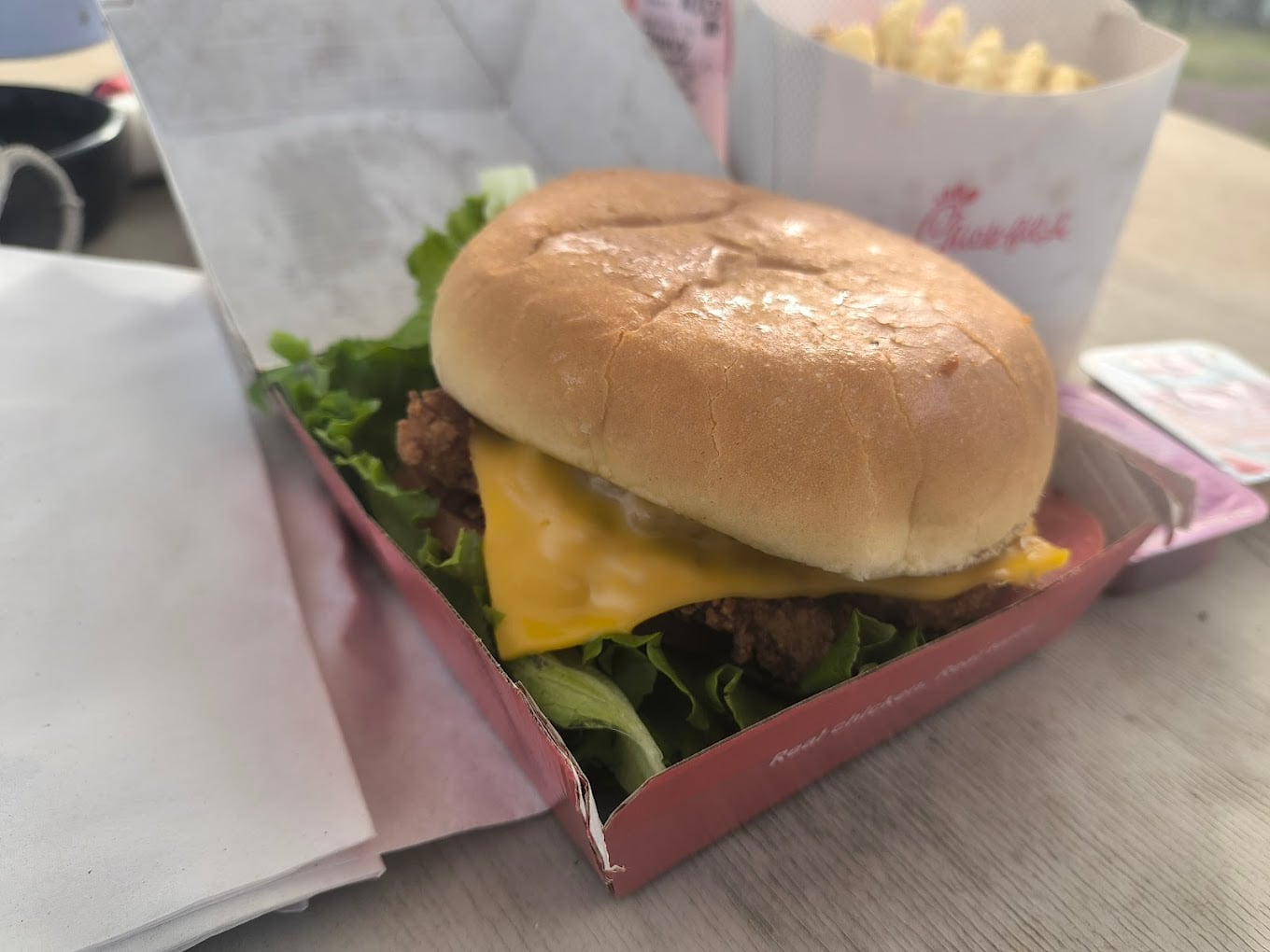 A chicken sandwich with lettuce and a slice of cheese in an open box sits on a table—just like you'd find at the favorite fast-food chains by state—alongside crinkle-cut fries and a sauce pack in the background.
