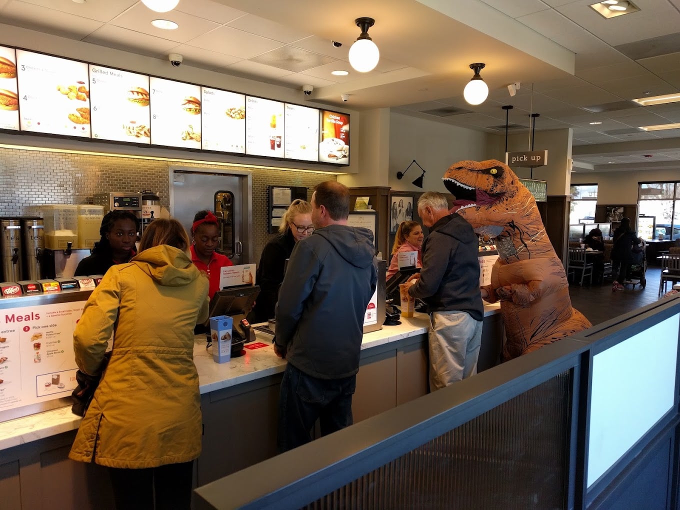 Several people order food at a fast-food counter, including one person in an inflatable dinosaur costume. Restaurant staff assist customers under menu boards, creating a fun scene reminiscent of favorite fast-food chains by state.