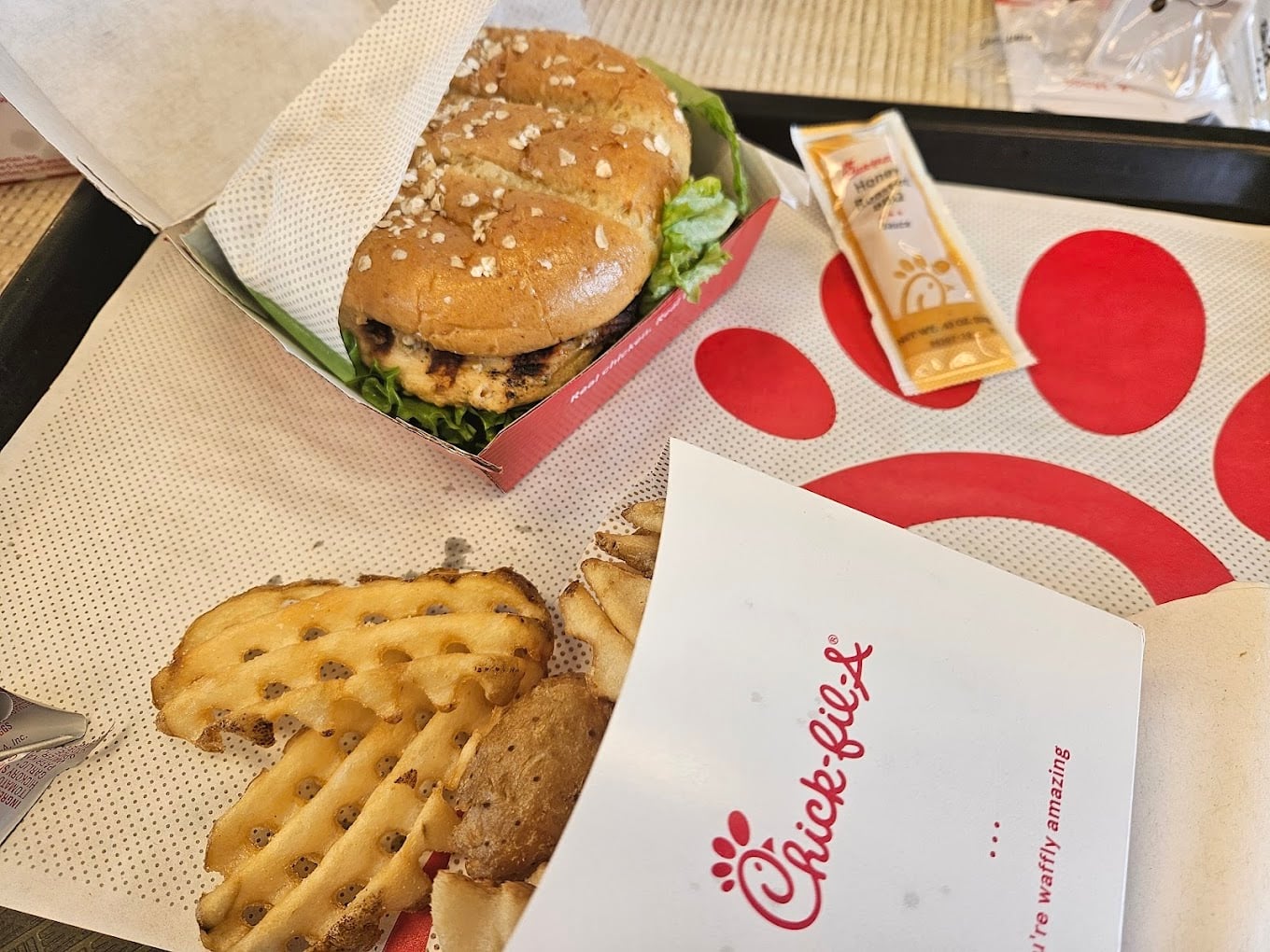 A Chick-fil-A meal with a grilled chicken sandwich in a box, waffle fries spilling from a Chick-fil-A container, and a packet of honey roasted BBQ sauce on a tray—one of the favorite fast-food chains by state.