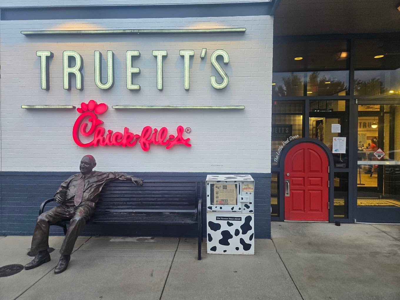 A bronze statue of a man sits on a bench outside Truett's Chick-fil-A, one of the favorite fast-food chains by state. Nearby is a cow-patterned donation box, a small red door by the entrance, and neon signage above.
