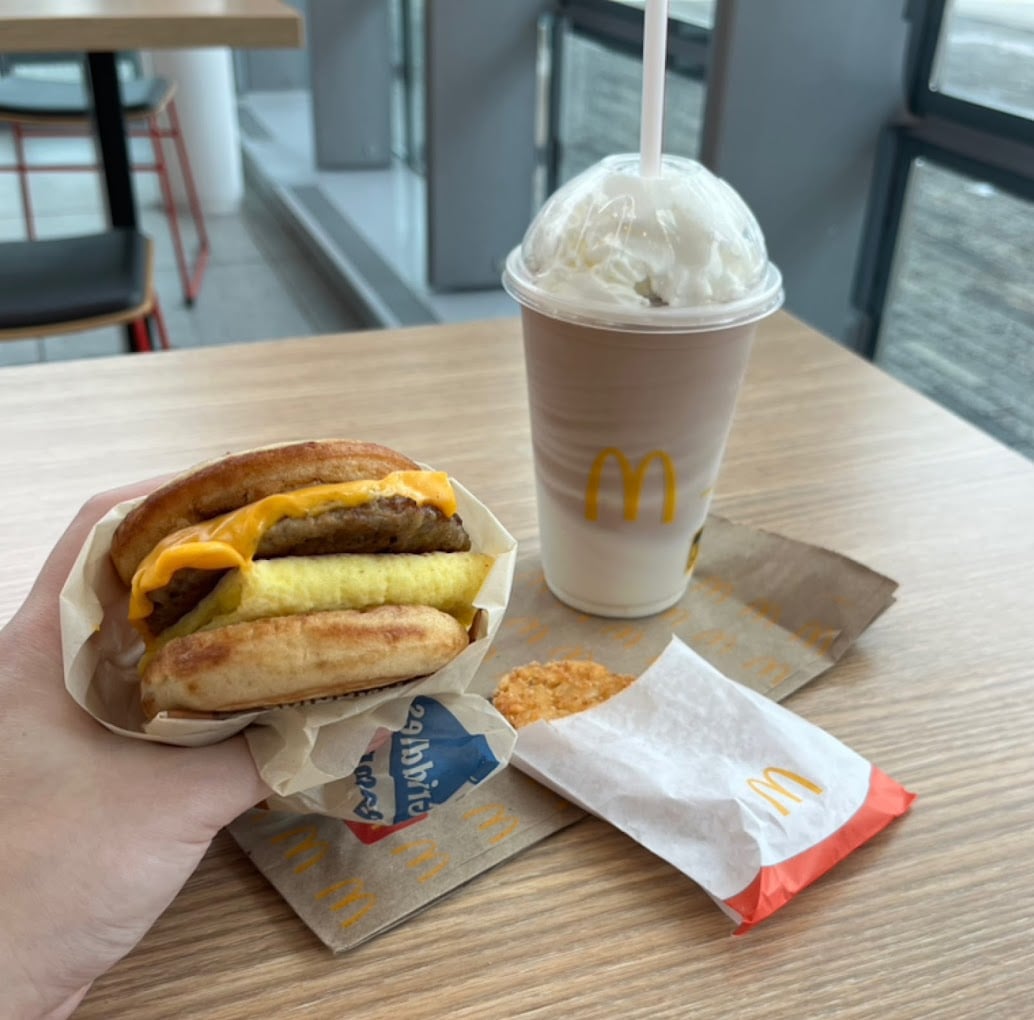 A hand holds a McDonald's breakfast sandwich with egg, cheese, and sausage on an English muffin. On the table are a hash brown in a paper sleeve and a vanilla milkshake—classic fare from one of the favorite fast-food chains by state.
