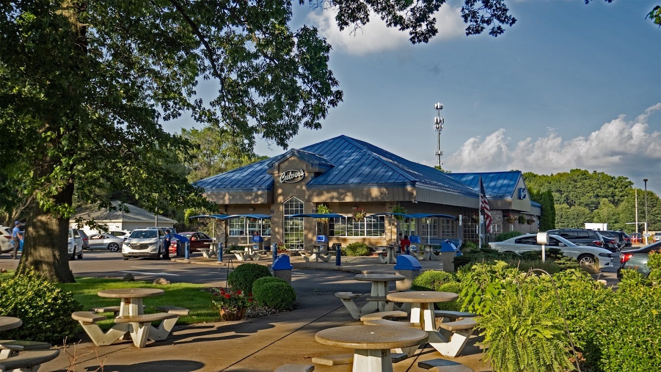 A roadside rest area features a building with a blue roof, surrounded by trees and greenery. Picnic tables dot the foreground, while parked cars—perhaps travelers seeking favorite fast-food chains by state—gather near the building under a partly cloudy sky.