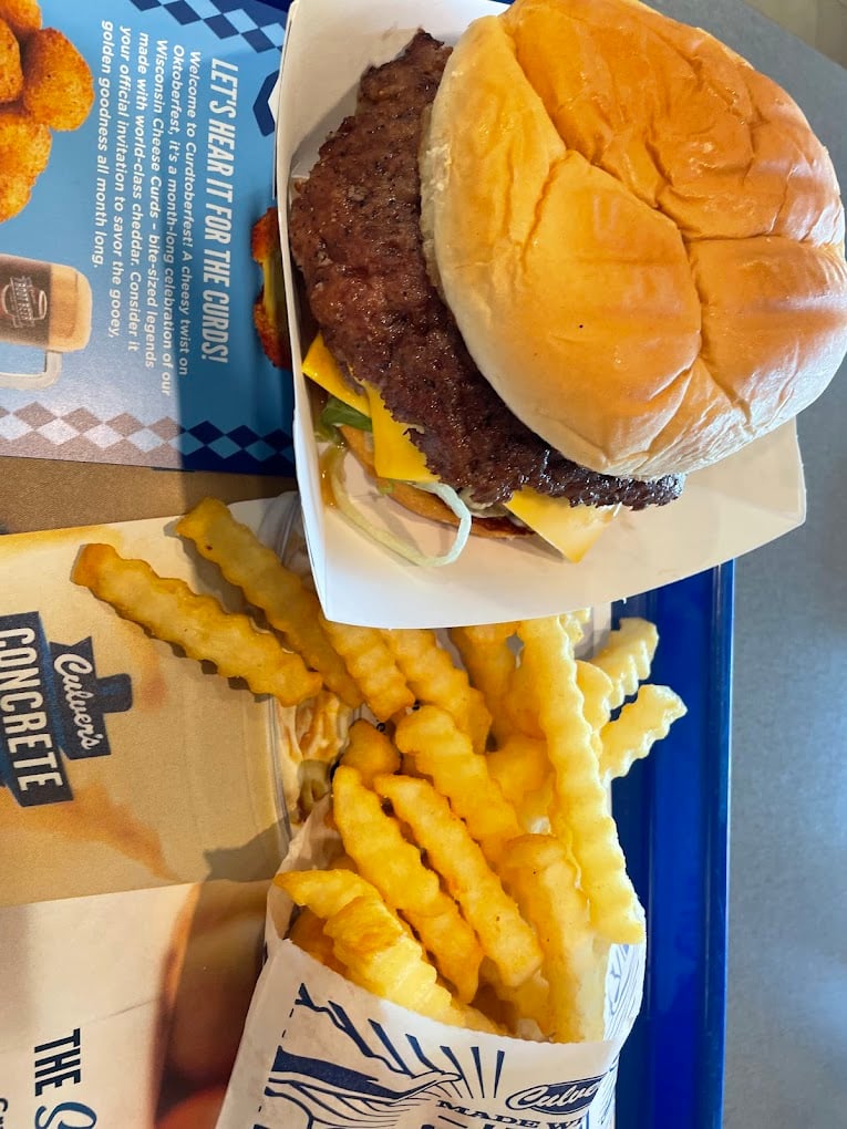 A cheeseburger with lettuce, tomato, and onions in a paper tray sits beside crinkle-cut fries on a blue tray with a Culver’s menu underneath—no wonder it’s among the favorite fast-food chains by state.