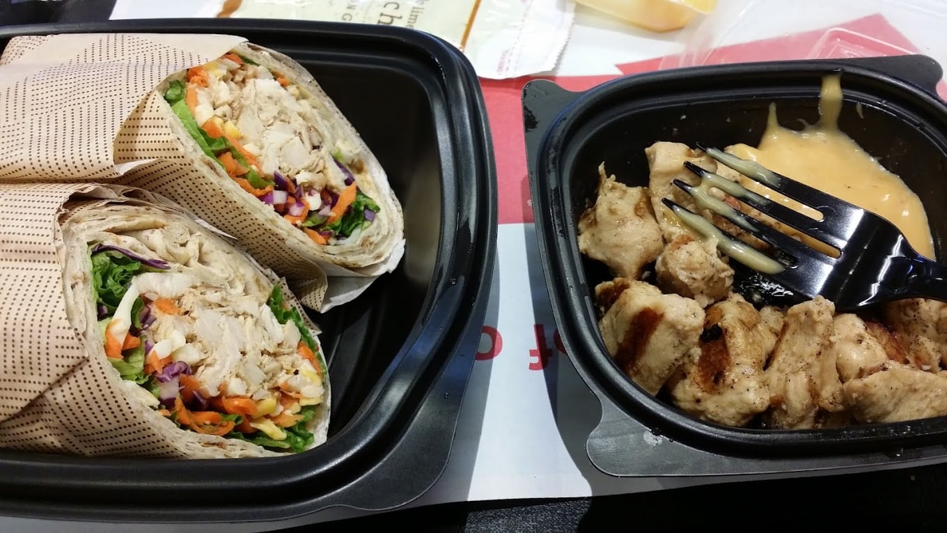 Two containers of food: one holds a wrap cut in half with shredded chicken, lettuce, carrots, and cabbage; the other features grilled chicken pieces with creamy dipping sauce—a meal you might find at favorite fast-food chains by state.