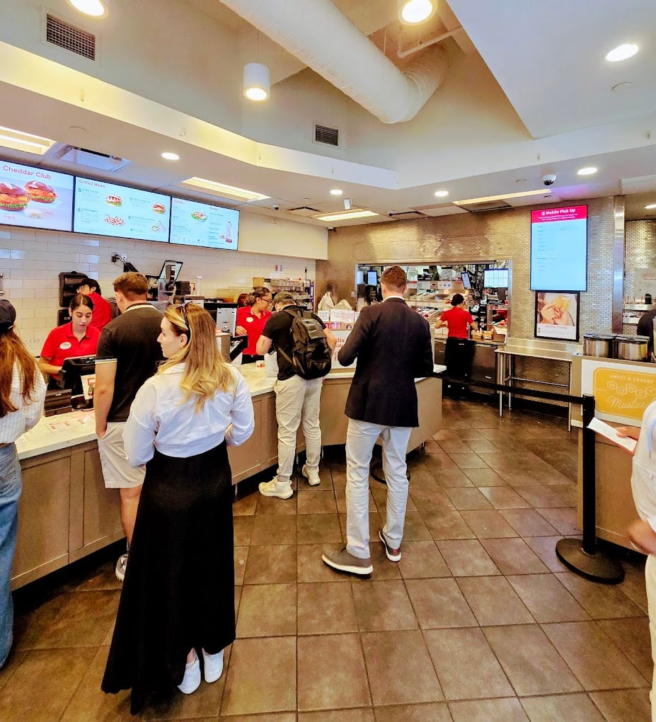 Customers stand in line at a fast-food restaurant counter, ordering food from employees in red shirts. Digital menu boards hang above, showcasing one of the favorite fast-food chains by state amid tiled floors and bright lighting.
