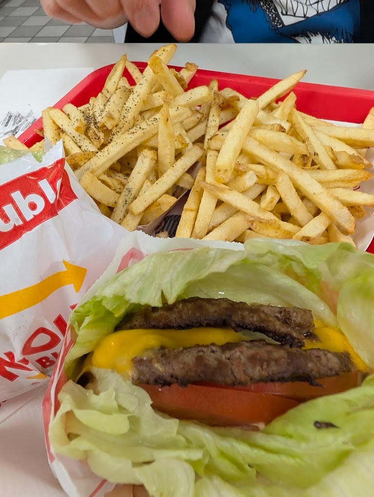 A meal on a red tray from one of the favorite fast-food chains by state features a cheeseburger wrapped in lettuce with tomato and two beef patties, next to a pile of thin, seasoned French fries. A person's hand is visible at the top.