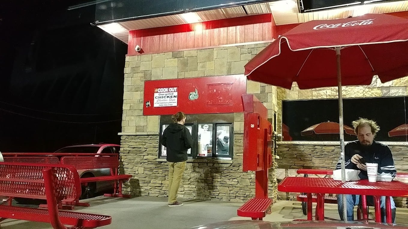A person stands at an outdoor order window under bright lights at a restaurant with red benches and umbrellas, reminiscent of the favorite fast-food chains by state; another person sits at a table eating and drinking at night.
