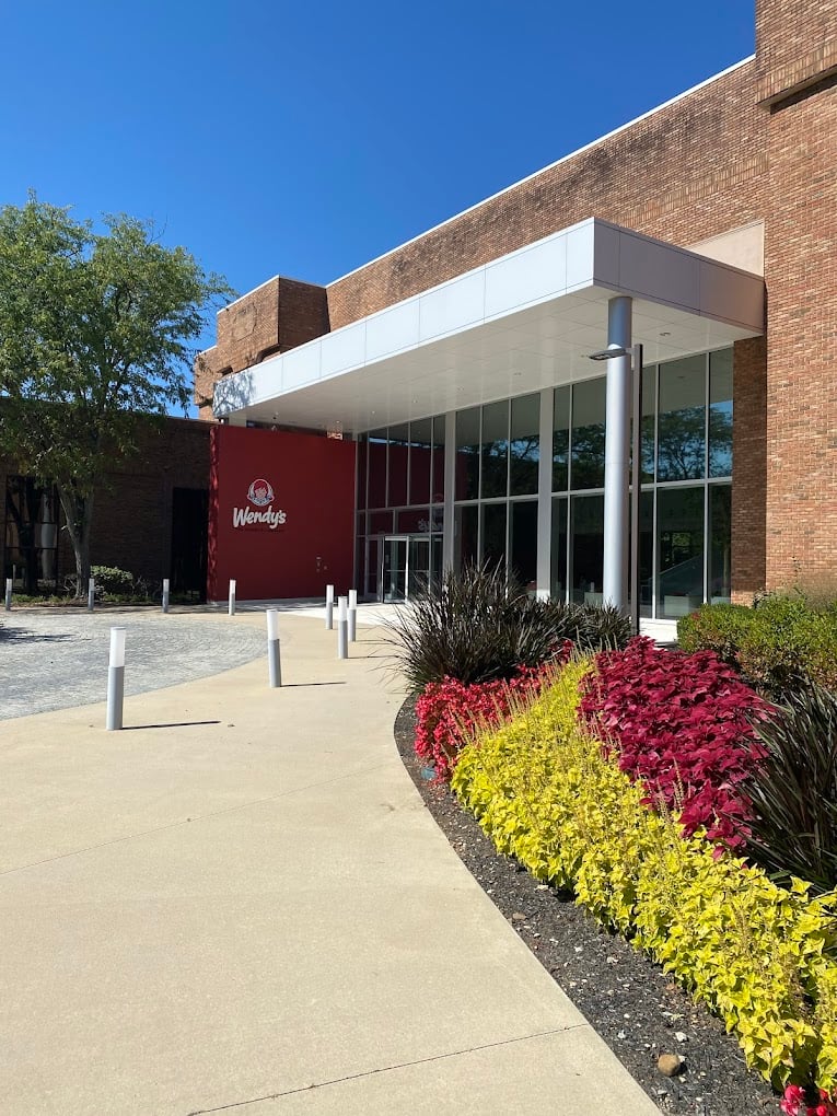 A Wendy’s restaurant with a modern glass entrance, red logo wall, and flower beds shines under a clear blue sky. White bollards line the curved sidewalk—an inviting look for one of the favorite fast-food chains by state.