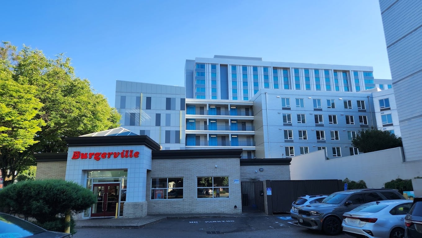 A Burgerville restaurant, one of the favorite fast-food chains by state, stands with its red sign in front of a modern multi-story building. Several cars are parked nearby, while green trees line the scene under a clear blue sky.