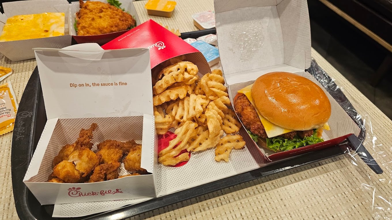A tray with Chick-fil-A food—waffle fries, a fried chicken sandwich with lettuce and cheese, and chicken nuggets. In the background are more food items and sauce packets from one of the favorite fast-food chains by state.