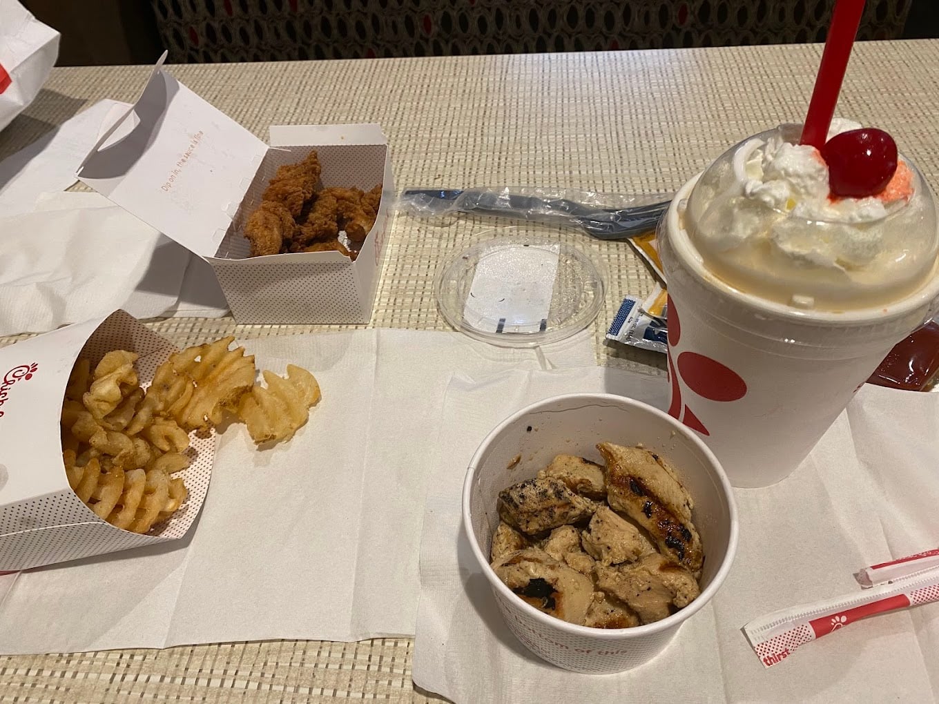 A meal inspired by favorite fast-food chains by state features waffle fries, chicken strips, grilled chicken pieces in a cup, a vanilla milkshake topped with whipped cream and a cherry, and wrapped utensils on the table.