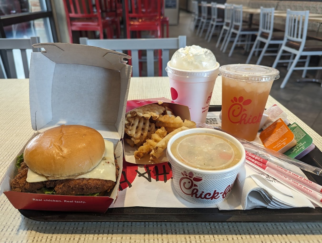 A Chick-fil-A meal tray featuring a chicken sandwich, waffle fries, chicken noodle soup, iced tea, a milkshake, and sauces sits on a table in one of the top favorite fast-food chains by state, with empty chairs in the background.