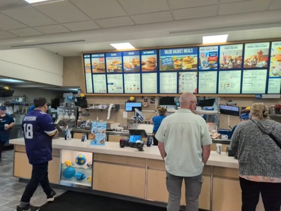 Customers stand at the counter of one of the favorite fast-food chains by state, looking at the menu boards above. Employees in blue uniforms prepare orders behind a clean, brightly lit counter.