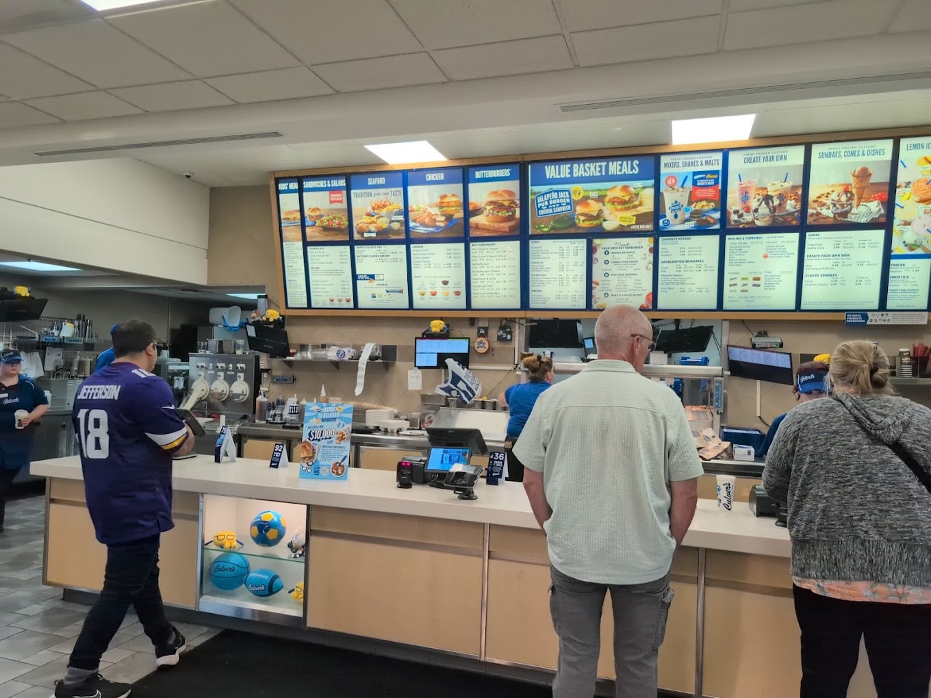 Customers stand at the counter of one of the favorite fast-food chains by state, looking at the menu boards above. Employees in blue uniforms prepare orders behind a clean, brightly lit counter.