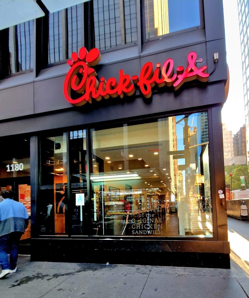 Street view of a Chick-fil-A restaurant—one of the favorite fast-food chains by state—with large glass windows, the red Chick-fil-A logo above the entrance, and a few people walking by on the sidewalk in an urban setting.