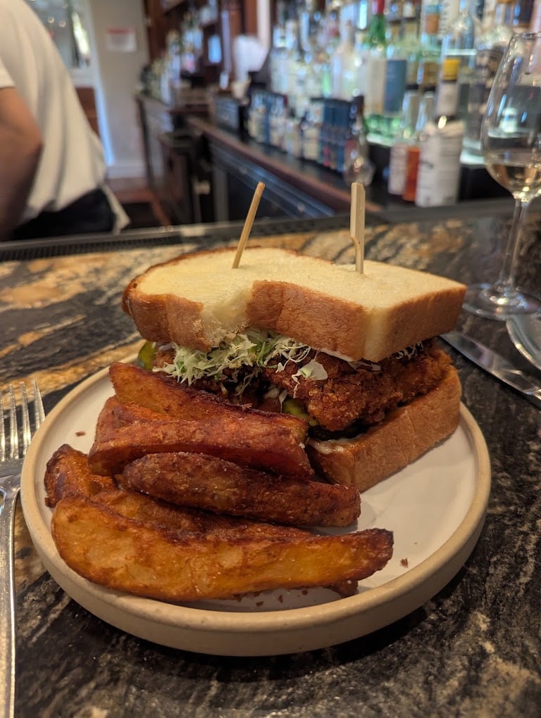 A sandwich with lettuce and fried filling on white bread is served on a plate with thick potato wedges. The plate sits on a marble bar counter, with bottles and a glass of white wine in the background.