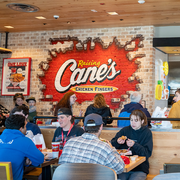 People are dining inside a Raising Cane’s restaurant, known as one of the favorite fast-food chains by state. The brick wall features a large Cane’s Chicken Fingers logo, and several groups enjoy meals in the lively, casual atmosphere.
