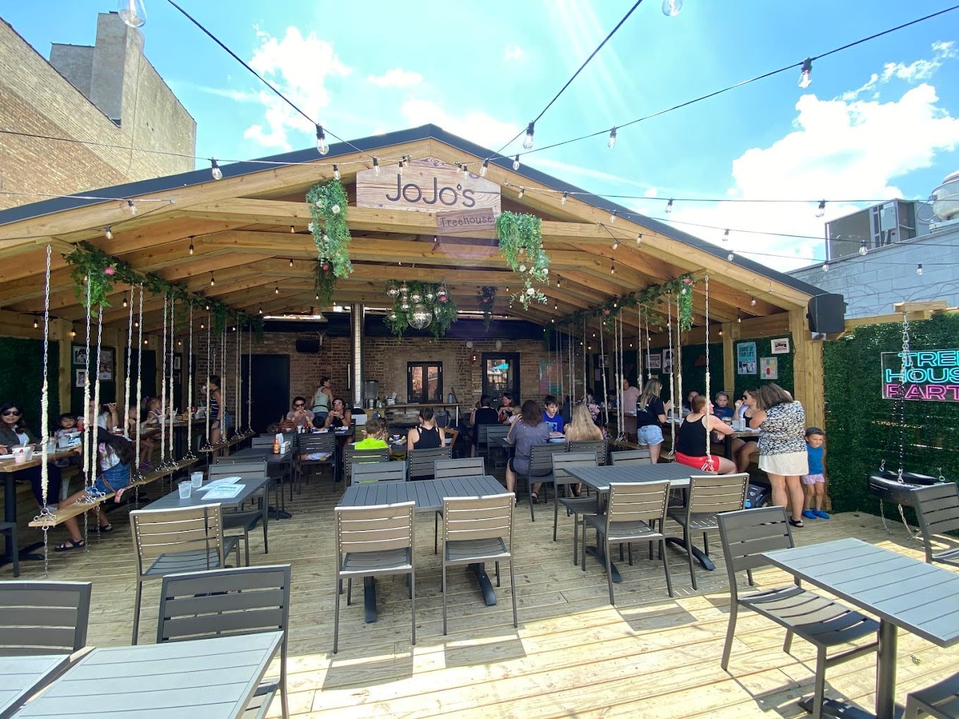 Outdoor patio at JoJo’s restaurant with tables and chairs, people dining, hanging lights, greenery decorations, and a wooden roof on a sunny day. The atmosphere is lively and welcoming.