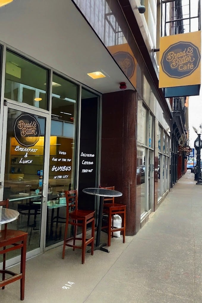 Street view of Bread & Butter Café with yellow signage, glass entrance, outdoor tables and chairs, and handwritten messages on the door advertising breakfast, lunch, catering, and delivery options.