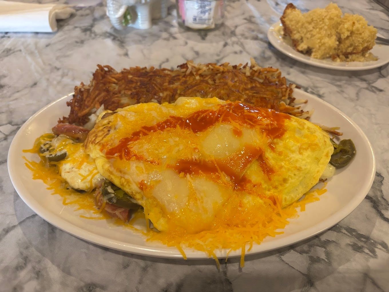 A plate with a cheesy omelette topped with hot sauce, served alongside crispy hash browns. In the background, there is a plate with a slice of crumbly cake on a marble table.