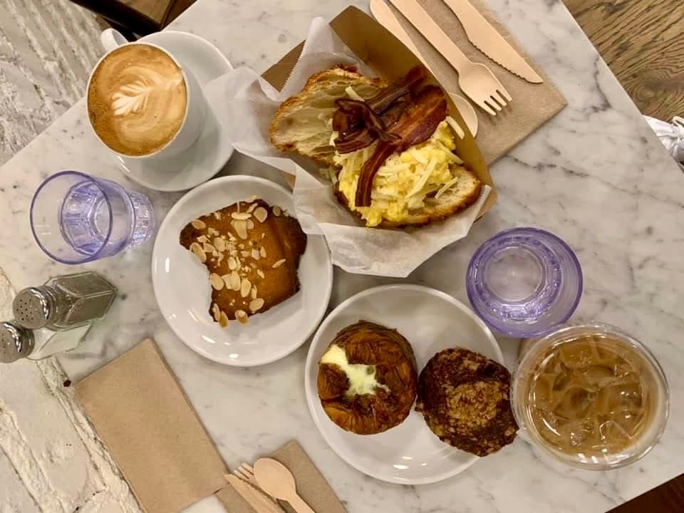 Breakfast spread on a marble table with a croissant sandwich, slice of almond cake, two pastries, the best hash browns, a latte, iced coffee, two glasses of water, wooden cutlery, napkins, and a salt shaker.