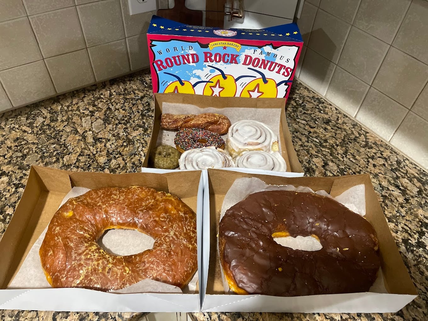 Two large donuts, one glazed and one chocolate-covered, sit in separate boxes on a granite counter. Behind them are assorted pastries and a colorful "Round Rock Donuts" box against a tiled backsplash.