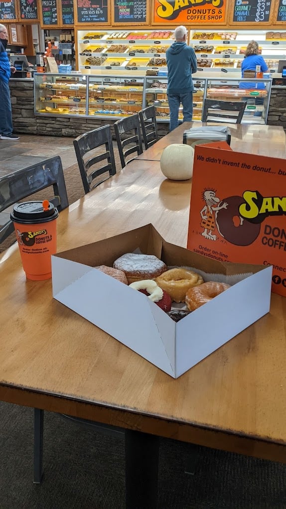 A box of assorted donuts, a drink cup, and an orange takeout bag sit on a wooden table in a donut shop with a display case and menu boards in the background.