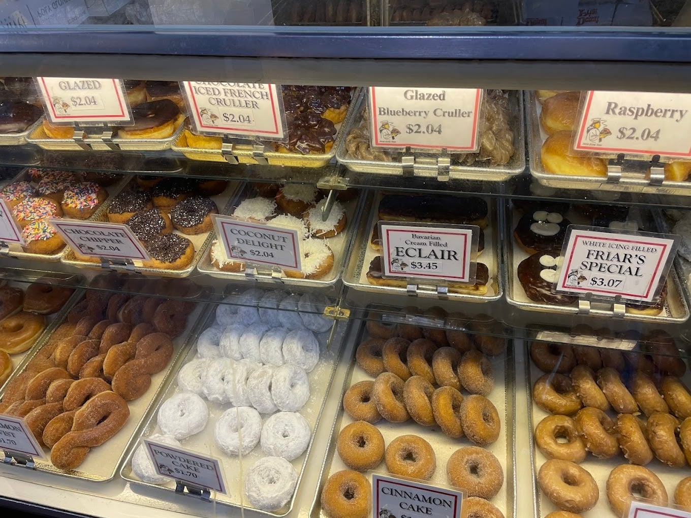 A display case filled with various donuts and pastries, including glazed, chocolate, coconut delight, eclairs, powdered sugar, and cinnamon cake donuts, each with price labels above or in front of them.