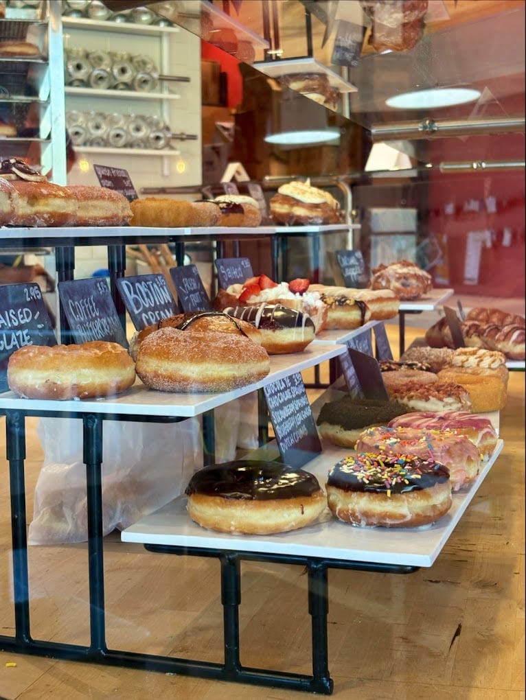 A bakery display case filled with assorted donuts, including ones with chocolate glaze, sprinkles, powdered sugar, and cream fillings, each labeled with small signs. Shelves and baking supplies are visible in the background.