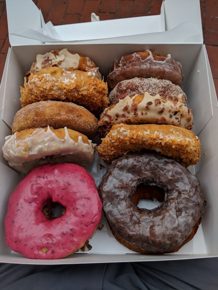A box of six assorted donuts, including ones with pink, white, and chocolate glaze, and others topped with coconut flakes and crumbled nuts, sitting on someone's lap.