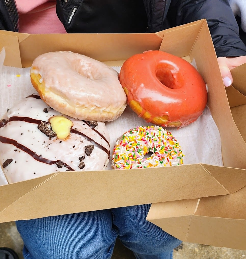 A person holds an open box containing four donuts: a glazed donut, a red frosted donut, a white frosted donut with chocolate drizzle and cookie pieces, and a white frosted donut with colorful sprinkles.