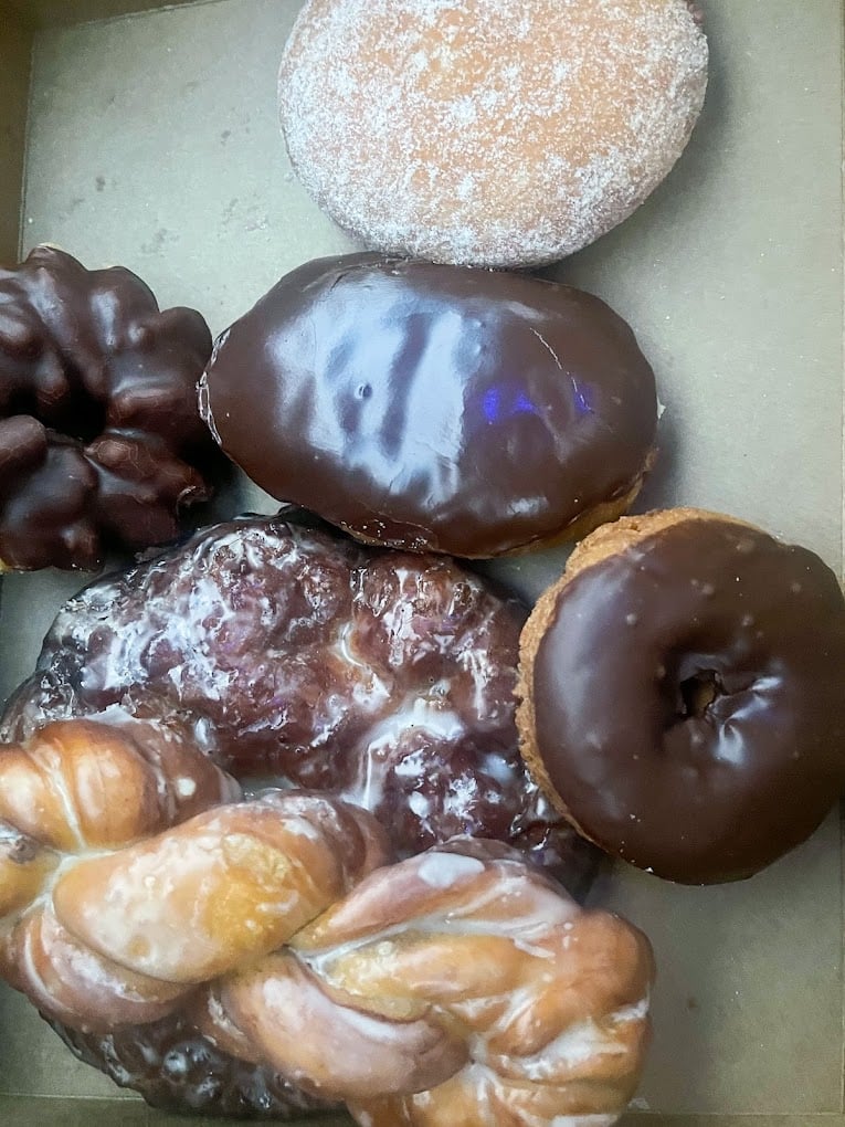 A box with six assorted donuts, including a chocolate-glazed donut, a glazed twist, an apple fritter, a powdered jelly donut, a chocolate cake donut, and a chocolate-covered round donut.